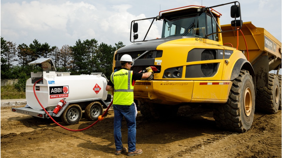 A picture of a construction worker wearing a safety vest and a white hard hat. The construction worker is refueling an off-road vehicle with a mobile and on-site fuel tank.